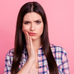 Brunette woman in a plaid shirt cringes in pain and touches her cheek due to tooth sensitivity