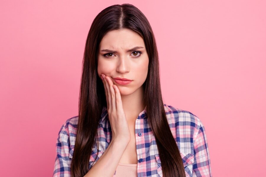 Brunette woman in a plaid shirt cringes in pain and touches her cheek due to tooth sensitivity