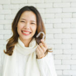 Asian woman in a cream sweater smiles against a white brick wall as she holds up her Invisalign aligners