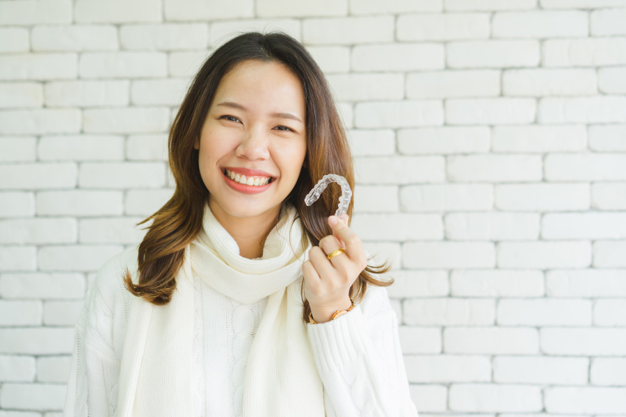 Asian woman in a cream sweater smiles against a white brick wall as she holds up her Invisalign aligners