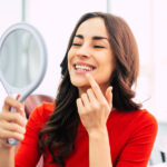 Brunette woman in a red blouse smiles and points to her smile after teeth whitening