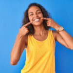 Brown woman in a yellow tank top smiles after professional teeth whitening and points to her teeth against a blue wall