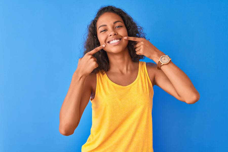 Brown woman in a yellow tank top smiles after professional teeth whitening and points to her teeth against a blue wall