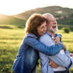 A middle-aged woman and man embrace while smiling outside in a grassy meadow as the sun sets