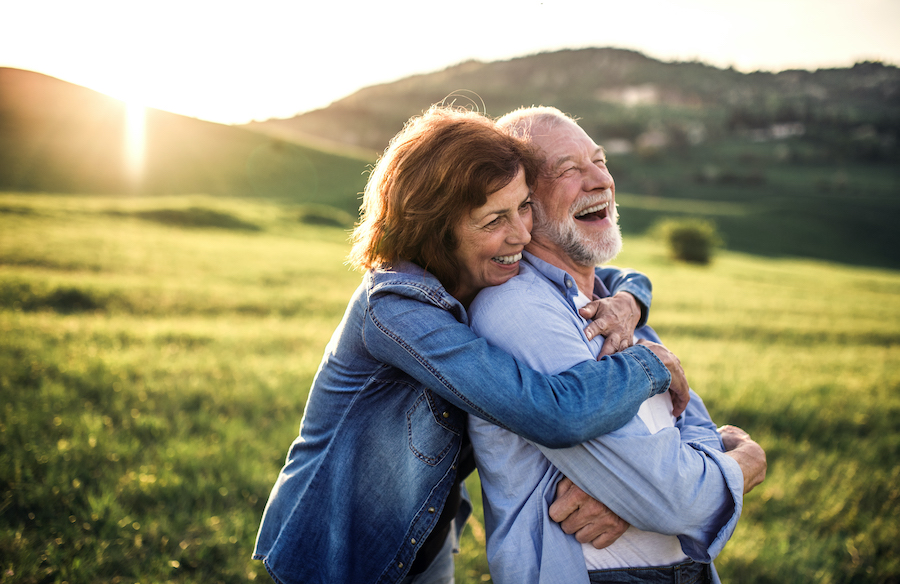A middle-aged woman and man embrace while smiling outside in a grassy meadow as the sun sets
