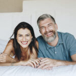 Middle-aged couple smile together on the bed with healthy, beautiful teeth